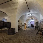 Gewölbekeller mit alten Bierfässern, Werkzeugen und Schildern auf Kopfsteinpflasterboden. Beleuchtung durch Deckenlampen.