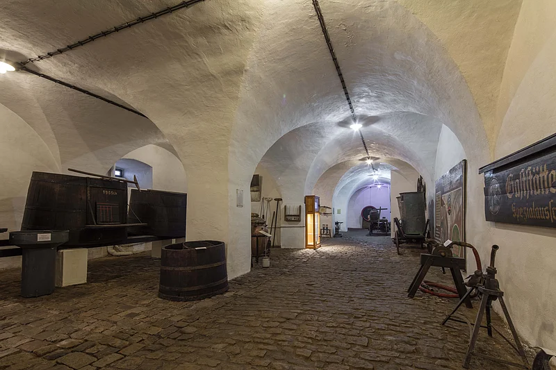 Gewölbekeller mit alten Bierfässern, Werkzeugen und Schildern auf Kopfsteinpflasterboden. Beleuchtung durch Deckenlampen.