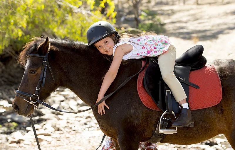 sweet beautiful young girl 7 or 8 years old riding pony horse hugging and smiling happy wearing safety jockey helmet posing outdoors on countryside in summer holiday